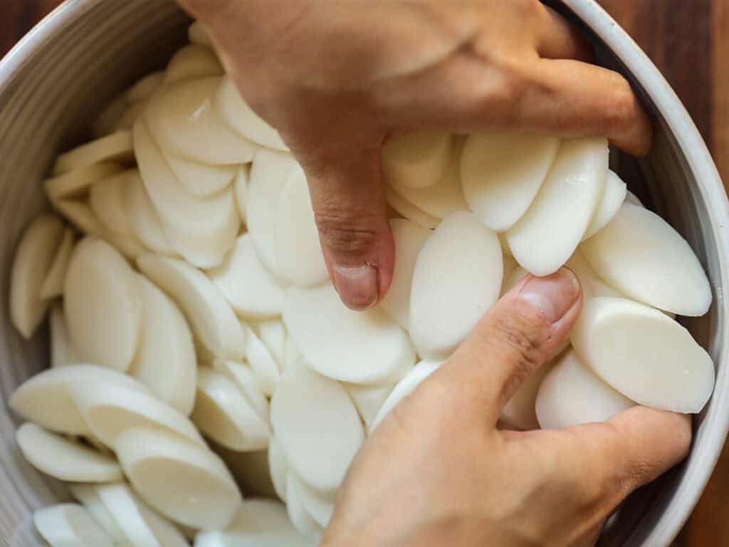 Hands holding oval-shaped, sliced rice cakes in a bowl, likely being prepared for cooking. The rice cakes are smooth and white, filling the bowl completely. The background is a wooden surface.